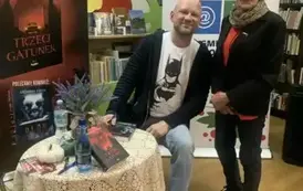 Starsza Pani stoi przy stoliku pisarza prosząc o podpis nowej książki. Z prawej strony stoi rolap z logiem biblioteki, a po lewej stronie rolap promujący nową książkę autora pt "Trzeci gatunek"