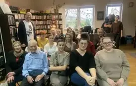 Uśmiechnięte twarze czytelnik&oacute;w siedzących na krzesłach, za nimi stoi uśmiechnięty pisarz, a z tyłu  jest czytelnik i bibliotekarka.