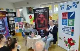 Przy stoliku z przodu siedzą pisarz i prowadząca spotkanie kierowniczka z teczką. Za nimi i po bokach stoją rolapy promujace książki autora, rolap z logo biblioteki i tablica z grafikami okładek książek pisarza.