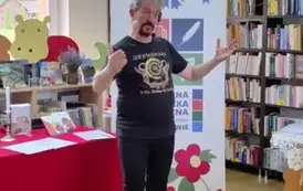 Pan Pilarski stoi z rozłożonymi rękami recytując wiersz. Za nim st&oacute;ł z czerwonym obrusem na kt&oacute;rym jest świeczka, kwiaty i książki  autora. Obok stoi rolap z logiem biblioteki.