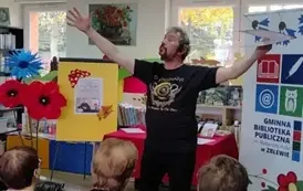 Pan Pilarski stoi z wysoko rozłożonymi rękami, w lewej ręce trzyma teczkę z tekstem. Recytuje wiersze. Za Nim rolap z logiem biblioteki, po środku st&oacute;ł, a po prawej metalowa tablica z plakatem dot. spotkania i duże kwiaty z krepy.