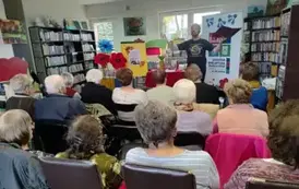 Widok z tyłu sali, krzesła zajęte przez gości. Z przodu Pan Pilarski stoi z rozłożonymi rękami, w kt&oacute;rych w jednej trzyma teczkę z tekstem. Za Nim rolap z logiem biblioteki, po środku st&oacute;ł z czerwonym obrusem, świeczką i kwiatami. A po lewej tablica.