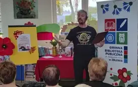 Pan Pilarski ma rozłożone ręce, w jednej trzyma tekst w teczce. Z przodu stoi st&oacute;ł z czerwonym obrusem, świeczką i kwiatami. Po prawej stronie stoi rolap z logiem biblioteki, a po lewej tablica z plakatem dot. spotkania i duże kwiaty z krepy.