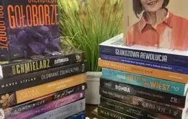 Na stole leżą książki ułożone pionowo,na nich jeszcze stoją dwie książki z widocznymi okładkami, przy nich leżą dynie dekoracyjne. W tle jesienny pejzaż.