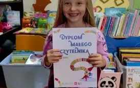 Uśmiechnięta dziewczynka w rozpuszczonych blond włosach siedzi na krzesłku w bibliotece w Pinczynie i pokazuje sw&oacute;j dyplom Małego Czytelnika