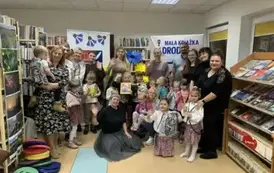 Grupa rodzic&oacute;w wraz z dziećmi z Panią prowadzącą warsztaty. Jedna dziewczynka w środku trzyma książkę., a niekt&oacute;rzy rodzice mają wyciągnięte rękę w geście machania.