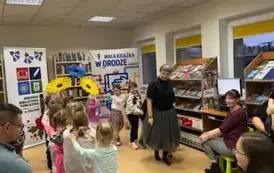 Dzieci idą wężykiem jeden za drugim za Panią Małgosią trzymając się za ramiona. Po bokach na krzesłach siedzą rodzice.