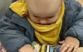 Malutki chłopiec w blond włoskach stoi przy p&oacute;łce z bajeczkami i patrzy na książeczki.