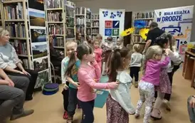 Dzieci idą w k&oacute;łeczku za Panią Małgosią jeden za drugim. Po bokach na krzesłach siedzą rodzice.