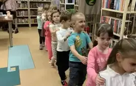 Dzieci idą w koło biblioteki jeden za drugim trzymając się za ramię.