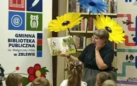 Dzieci partrzą na Panią Małgosię, kt&oacute;ra czyta im bajkę z książką skierowaną do nich, ma zszokowaną minę i rekę położoną na policzku. Za nią stoją kwiaty z krepy i po lewej stronie widac rolap z logiem biblioteki.