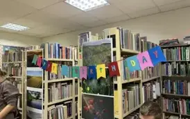 Na regałach z książkami wisi linka z przyczepionymi kolorowymi kartkami na kt&oacute;rych są literki składające napis Happy birth day.