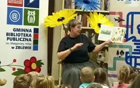 Dzieci siedzą na siedziskach i patrzą w stronę Pani Małgosi, kt&oacute;ra czyta im bajkę pokazując wnętrze książki. Za Nią stoją kwiaty z krepy, po lewej rolap z logiem biblioteki , a po prawej rolap " Mała książka w drodze"