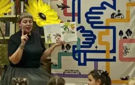 Dzieci siedzą na siedziskach i patrzą w stronę Pani Małgosi, kt&oacute;ra ma otwarte książkę pokazując jej wnętrze i ma podniesione palec wskazujacy. Z tyłu stoją kwiaty z krepy i po prawej rolap promujący "Mała książka w drodze"