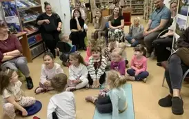 Dzieci siedzą na siedziskach w bibliotece, a za nimi z tyłu na krzesłach siedzą uśmiechnięci rodzice.