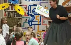 Dzieci siedzą na siedziskach,a przy nich stoi z otwartą książką Pani Małgosia lekko pochylona i z miną m&oacute;wiącą coś ciekawego. Z przodu widać kwiaty z krepy rolap " Mała książka w drodze"