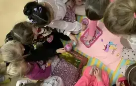 Dziewczynki siedzą wokoło dywanika na kt&oacute;rym są plastikowe talerzyki, kubeczki i dzbanek i biorą te przedmioty do rączek.