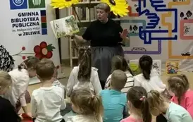 Dzieci siedzą na siedziskach i patrzą na Panią Małgorzatę, kt&oacute;ra z przodu czyta im bajkę pokazując wnętrze książki. Z tyłu są kwiaty z krepy, a po bokach rolapy z logiem biblioteki i rolap "Mała książka w drodze"