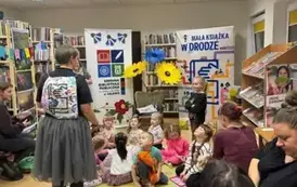 Dzieci siedzą na dywaniku ,a przy nich stoi Pani Małgosia, obok siedzą na krzesłach rodzice. Z przodu widać po bokach rolapy i kwiaty po środku z krepy.