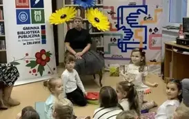 Dzieci siedzą na siedziskach, z przodu siedzi na krzesłku Pani Małgosia. Za Nią stoją trzy kwiaty zrobione z krepy, dwa ż&oacute;łte i jeden niebieski. Po prawej stronie stoi rolap "Mała książka w drodze",a po lewej rolap z logiem biblioteki.