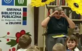 Dzieci siedzą na siedziskach i się rozglądają wokoło. Z przodu Pani Małgosia ma położone książkę na kolanach, a dłonie ułożone przy oczach w kształcie okular&oacute;w. Za Nią stoją trzy sztuczne kwiaty z krepy, po lewej rolap z logiem biblioteki.