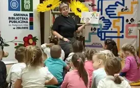 Dzieci siedzą na siedziskach, a z przodu Pani Małgorzata czyta im bajkę. Za Nią stoją trzy kwiaty zrobione z krepy, a po bokach stoją rolapy- po lewej rolap biblioteki a po prawej rolap Mała Książka w drodze.