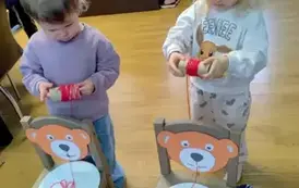 Dwie dziewczynki stoją za krzesełkami na kt&oacute;rych siedzeniach są położone papierowe talerzyki z rozwiniętą wł&oacute;czką. Na oparciach są naklejone głowy misia i dzieci stojąc z tyłu nawiajają wł&oacute;czkę na rolkę wizualizując jedzenie makaronu.