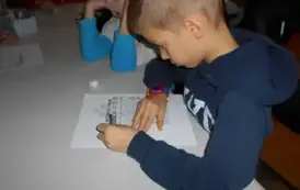 Chłopiec w granatowej bluzie przy stole uzupełnia misiową kartę pracy, obok widać dziewczynkę, kt&oacute;ra ma opartą głowę na łokciach.