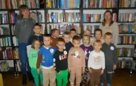 Grupa dzieci wraz ze swoją Panią i z Panią bibliotekarką stoją przy p&oacute;łkach z ksążkami do zdjęcia.