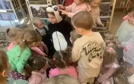 Na dywanie siedzi kobieta przebrana za misia Pandę, otoczona jest dziećmi, kt&oacute;rzy ją dotykają i chcą się przytulić.