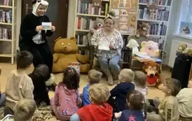 Dzieci siedzą na dywanie, a z przodu siedzi bibliotekarka trzymając książeczkę i pokazując ją, a z  lewej strony siedzi kobieta przebrana za misia pandę i pokazuje im broszurę o segregowaniu śmieci.