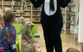 Dzieci siedzą na dywanie, a przy nich stoi kobieta przebrana za misia Pandę, ma rozłożone ręce w bok.