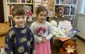 Przy fotelu z misiami stoi chłopiec i dziewczynka, obok na podłodze siedzi brązowy miś, a za nimi stoi korkowa tablica  z grafikami dot. święta misia.