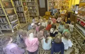 Dzieci siedzą na dywanie i patrzą do przodu na bibliotekarkę, kt&oacute;ra im opowaida o święcie Misia. Obok bibliotekarki siedzi duży brążowy miś, a po prawej na fotelu leżą małe misie i bajki do czytania. Na wprost stoi tablica przystrojna w grafikę o misiach