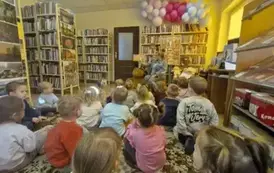 Dzieci siedzą na dywanie i patrzą do przodu jak bibliotekarka czyta im bajkę. Na szafie z książkami u g&oacute;ry leżą balony.