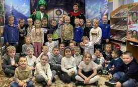 Spora grupa dzieci stoi razem z dwoma elfami. Jeden z elf&oacute;w ma buzię otwartą i wyciągnięte dwa place do g&oacute;ry.  W tle pok&oacute;j ,a po bokach zaśnieżony krajobraz z choinkami.