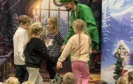 Z przodu zielony elf tańczy w k&oacute;łeczku z trzema dziewczynkami i dw&oacute;jką chłopc&oacute;w.