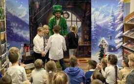 Z przodu zielony elf tańczy w k&oacute;łeczku z dwiema dziewczynkami i dwoma chłopcami.