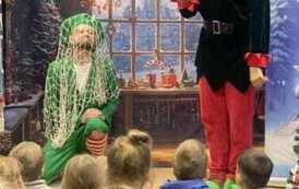 Po środku stoi elf z wyciągniętą ręką do g&oacute;ry z palcem wskazującym, a obok siedzi drugi elf z zarzuconą siatką na sobie. W tle pok&oacute;j z choinką.