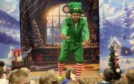 Na środku przed dziećmi stoi elf ubrany cały na zielono i jest pochylony i ma wyciągnięte ręce w kierunku dzieci. W tle pok&oacute;j z choinką, a po bokach zaśnieżone choinki.