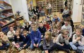 Na dywanie siedzi duża grupa dzieci, a z tyłu na krzesłach siedzą nauczyciele.