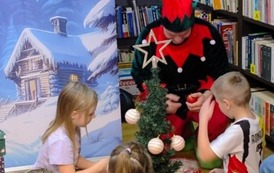 Z przodu przy małej choince siedzi elf i dwoje dzieci, chłopiec i dziewczynka i ubierają choinkę w bombki. W tle zaśnieżone choinki z drewnianą zaśnieżoną chatą.