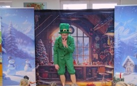 Po środku stoi pochylony elf i ma przyłożony palec do ust. W tle pok&oacute;j z choinką , a po bokach zaśnieżone choinki.