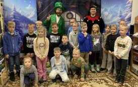 Na tle pokoju z choinką  i z bocznymi zaśnieżonymi choinkami stoją dzieci w grupie razem z dwoma elfami.