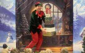 Na tle pokoju z choinką elf ubrany w czerwone spodnie i zieloną bluzkę z czerwonym kołnierzem wnosi na środek 7 karton&oacute;w z przezentami. 