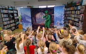 Na tle pokoju z choinką stoi pochylony elf ubrany na zielono, kt&oacute;ry pokazuje dzieciom ruchy do tańca. Dzieci stoją i naśladują elfa podnoszać ręce do g&oacute;ry.