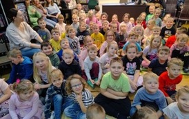 Na dywanie siedzi spora grupa dzieci, po bokach na krzesłach siedzą nauczyciele.