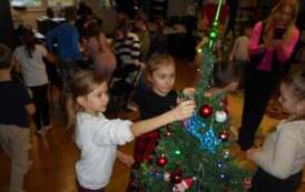 Dwie dziewczynki zawieszają na choinkę dekoracje, w tle są inne dzieci i ich Pani, kt&oacute;ra robi zdjęcia. 
