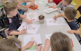 Dzieci siedzą przy białym stole i kolorują na swoich kartkach choinki.
