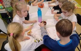 Dzieci siedzą przy białym stole i kolorują na swoich kartkach choinki według podanego kodu. Na stole stoi nap&oacute;j  w plastikowej butelce. 